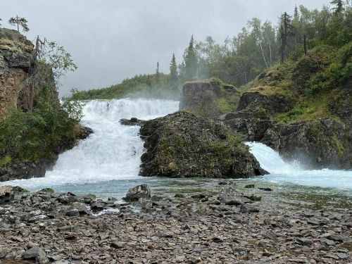 A waterfall cascades over rocky terrain, surrounded by lush greenery and mist under a cloudy sky.