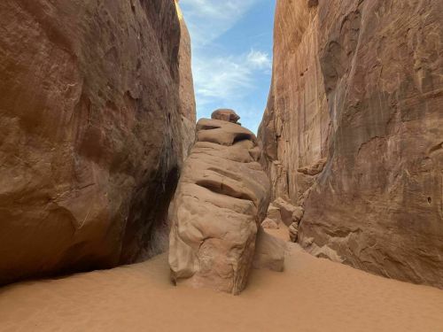A narrow canyon with towering rock walls and a large, smooth boulder in the center, surrounded by sandy ground.