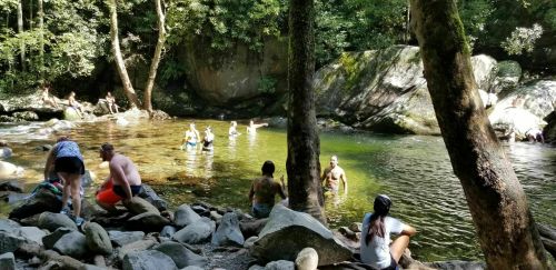 People enjoying a sunny day by a clear river, swimming and relaxing among rocks and lush greenery.