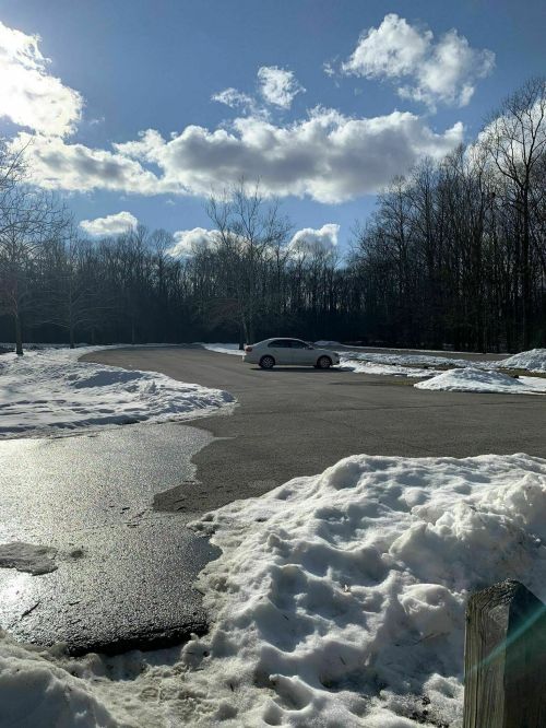 A snowy parking lot with a single car parked, surrounded by trees and a bright blue sky with fluffy clouds.