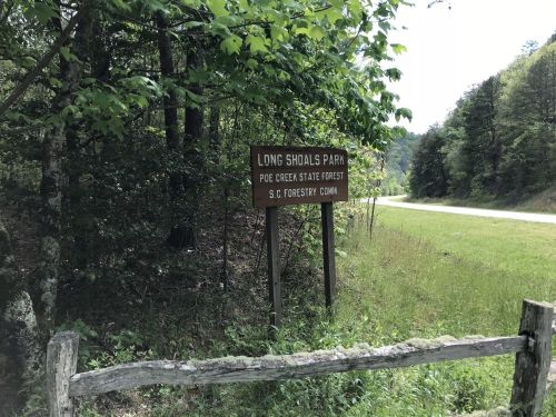 Sign for Long Shoals Park in Poe Creek State Forest, surrounded by greenery and a road in the background.