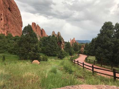 A winding path through lush greenery and red rock formations under a cloudy sky.