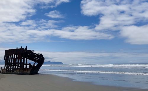 A weathered shipwreck on a sandy beach, with waves and a cloudy blue sky in the background.
