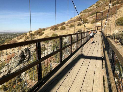 A person runs on a wooden suspension bridge with a mountainous landscape and valley view in the background.