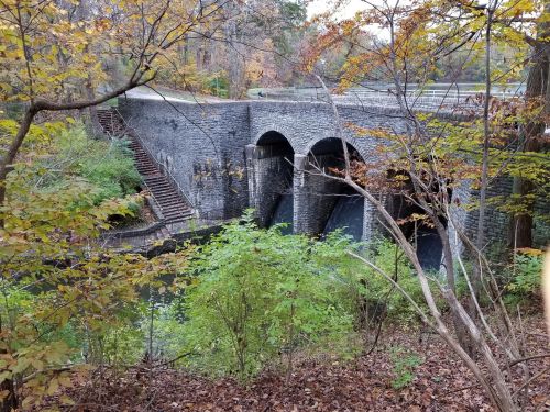 A stone bridge with three arches over a calm waterway, surrounded by autumn foliage and trees.