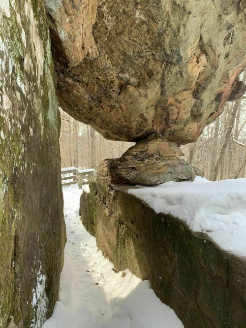 A large boulder balanced on a rock formation, with a snowy path and trees in the background.