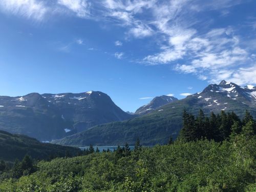 A scenic view of mountains and a lake, surrounded by lush greenery and a clear blue sky with scattered clouds.