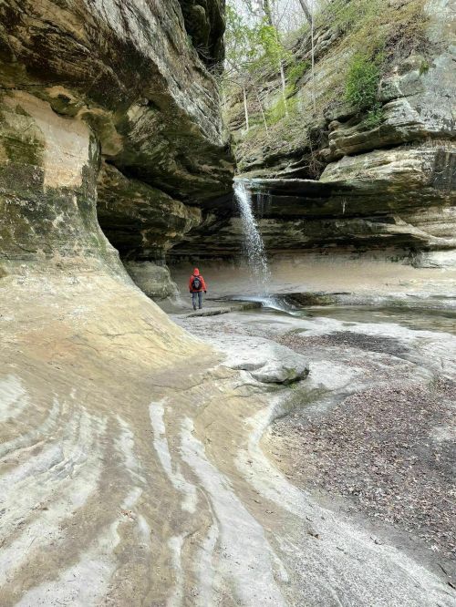 A person stands in a rocky canyon with a small waterfall cascading nearby, surrounded by lush greenery.