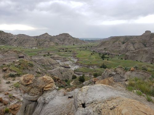 A panoramic view of rugged hills and valleys under a cloudy sky, with green patches of grass and a winding stream.