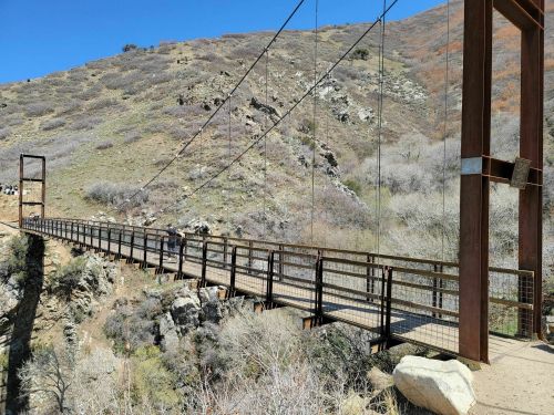 A suspension bridge spans a rocky canyon, surrounded by hills and sparse vegetation under a clear blue sky.