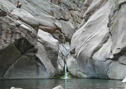 A rocky canyon with a small waterfall flowing into a pool, surrounded by large boulders and a person sitting above.
