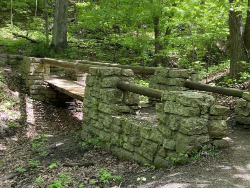 A rustic stone bridge with wooden railings, surrounded by lush green trees in a forested area.