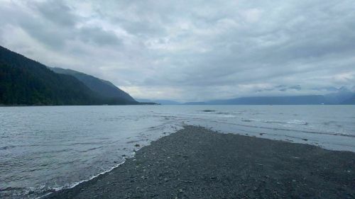 A rocky shoreline extends into a calm sea, surrounded by mountains under a cloudy sky.