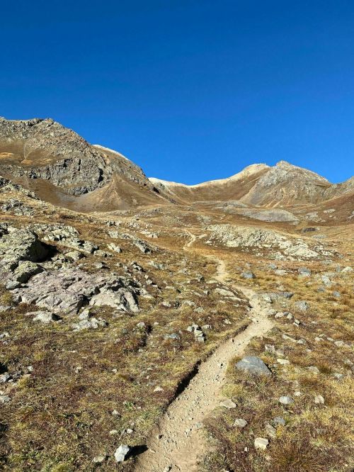 A winding dirt path leads through rocky terrain and golden grass, framed by rugged mountains under a clear blue sky.