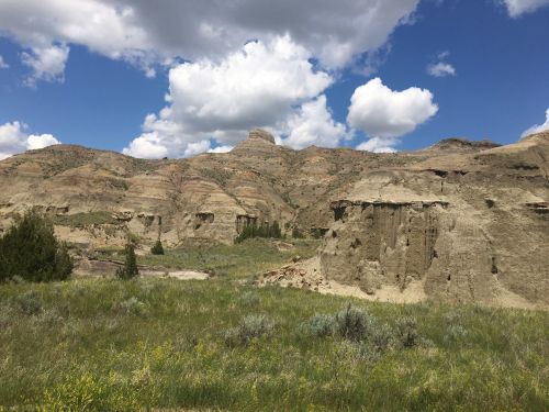 A scenic landscape featuring layered rock formations under a blue sky with fluffy clouds and green vegetation in the foreground.
