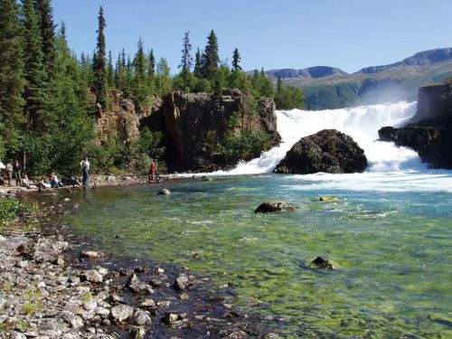 A scenic waterfall surrounded by lush greenery and rocky terrain, with people enjoying the view by the water's edge.