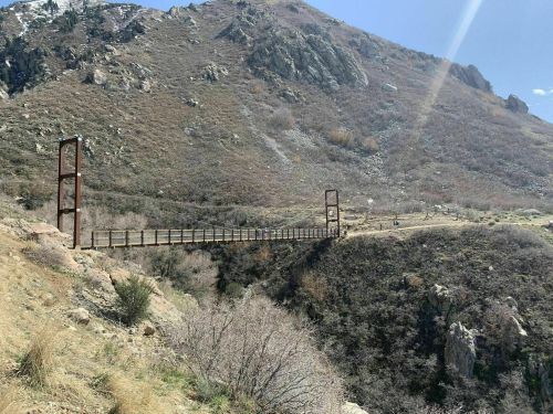 A suspension bridge spans a rocky canyon, surrounded by dry hills and sparse vegetation under a clear blue sky.