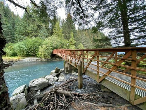 A wooden bridge spans a tranquil river, surrounded by lush green trees and rocky banks.