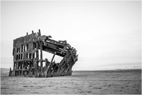 A weathered shipwreck stands on a sandy beach, partially submerged, against a soft, gray sky.