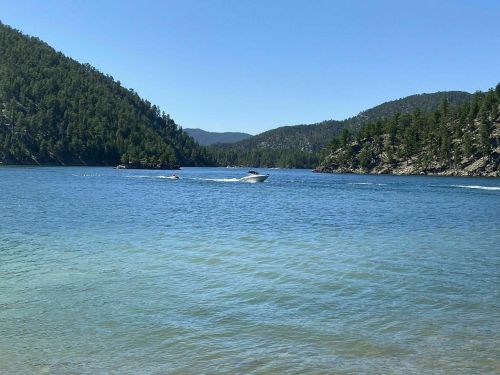 A boat speeds across a clear blue lake surrounded by green hills and trees under a bright blue sky.
