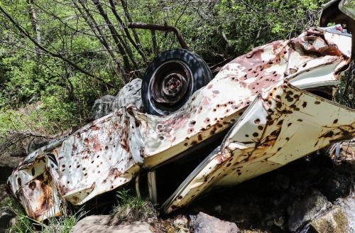 A rusted, overturned car partially submerged in a rocky stream, surrounded by greenery and trees.