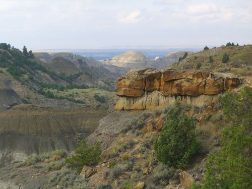 A rocky outcrop overlooks a vast, rugged landscape with rolling hills and sparse vegetation under a cloudy sky.