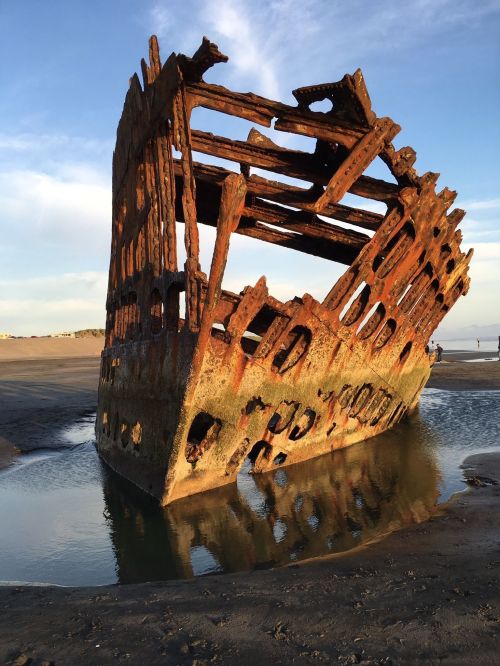 A weathered shipwreck partially submerged in shallow water on a sandy beach, with a clear blue sky in the background.