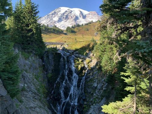 A waterfall cascades down rocky terrain, framed by lush green trees, with a snow-capped mountain in the background.