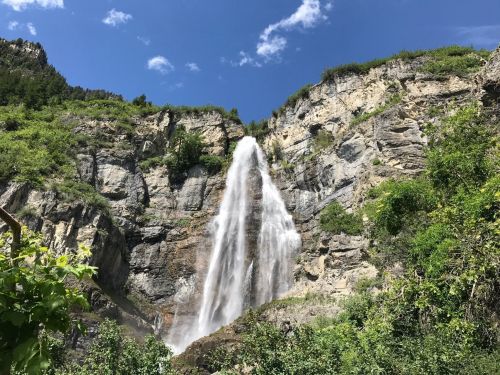 A tall waterfall cascading down a rocky cliff, surrounded by lush greenery and a clear blue sky.
