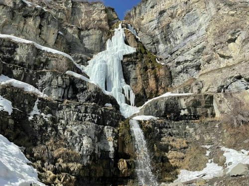 A frozen waterfall cascading down rocky cliffs, surrounded by snow and sparse vegetation under a clear blue sky.