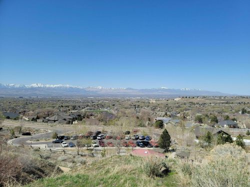 A panoramic view of a valley with houses, trees, and snow-capped mountains under a clear blue sky.