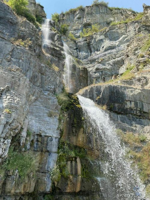 A cascading waterfall flows down a rocky cliff, surrounded by greenery and bright blue sky.
