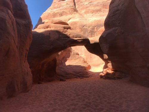 A sandy pathway leads through a natural rock arch in a red sandstone canyon under a clear blue sky.