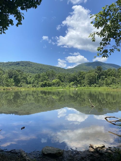 A serene lake reflects blue skies and clouds, surrounded by lush green mountains and trees.