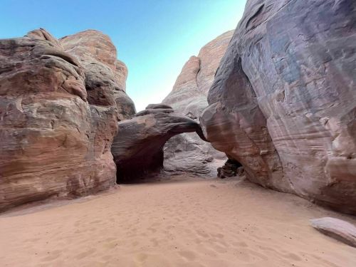 A sandy pathway leads through towering rock formations, featuring a natural arch in a desert landscape.