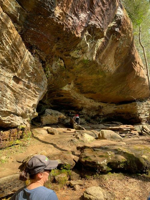 A person stands in front of a large rock formation with another person climbing above, surrounded by greenery.