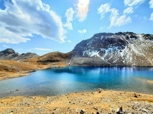 A serene mountain lake surrounded by rocky terrain and snow-capped peaks under a bright blue sky with fluffy clouds.