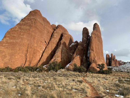 Red rock formations rise dramatically against a cloudy sky, with patches of snow and sparse vegetation in the foreground.