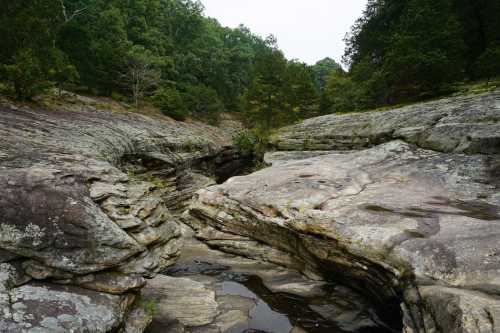 A rocky landscape with a narrow creek surrounded by lush green trees and a cloudy sky.