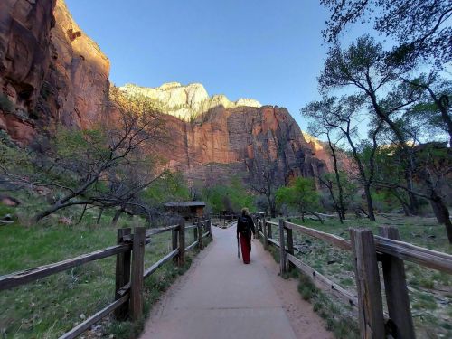 A person walks along a path in a canyon, surrounded by towering cliffs and greenery under a clear blue sky.