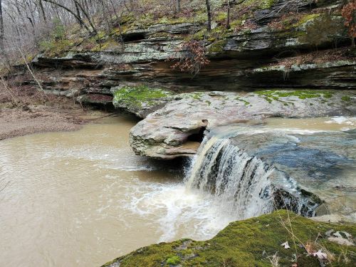 A small waterfall cascades over a rocky ledge into a muddy river, surrounded by moss-covered rocks and trees.