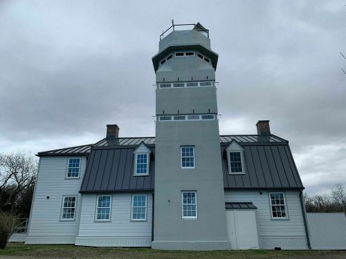 A tall, gray lighthouse-style building with a metal roof and multiple windows against a cloudy sky.