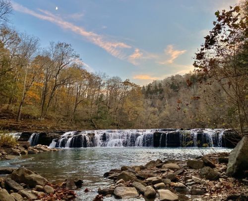 A serene waterfall cascades over rocks, surrounded by autumn trees under a colorful sky at dusk.