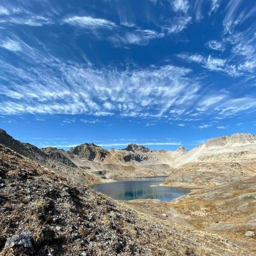 A serene mountain landscape featuring a calm lake surrounded by rugged terrain and a bright blue sky with wispy clouds.