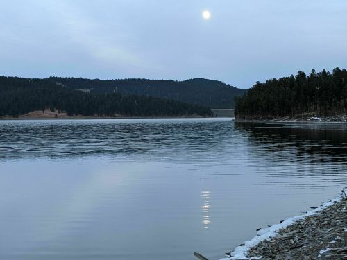 A calm lake reflects the moonlight, surrounded by forested hills under a cloudy sky.
