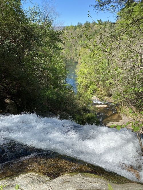A cascading waterfall flows over rocks, surrounded by lush greenery and a serene river in the background under a clear blue sky.