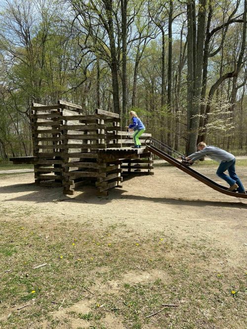 A child climbs a wooden play structure while another child pushes a slide in a forested park setting.