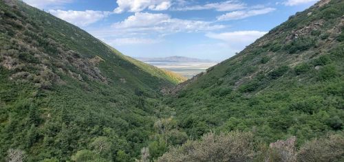 A lush green valley between two mountains, with a distant view of a lake and blue sky dotted with clouds.