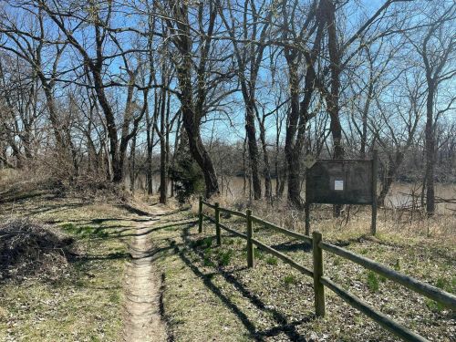 A winding dirt path beside a river, surrounded by bare trees and a wooden fence under a clear blue sky.