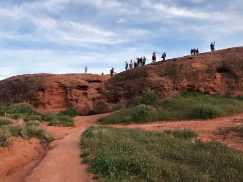 A group of people walking along a rocky ridge with red earth and green grass under a blue sky.